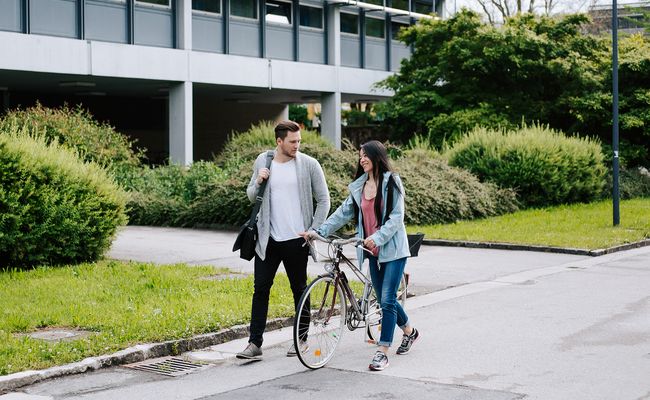 ein gehender Studierender und eine Rad schiebende Studierende neben dem Keplergebäude