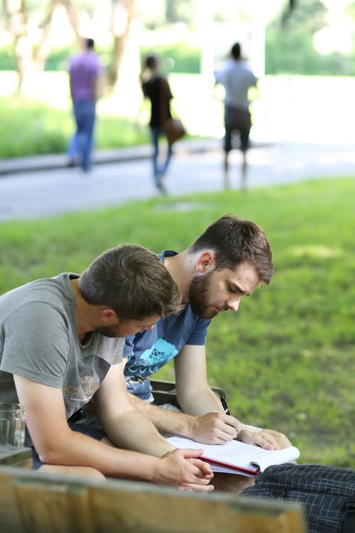 zwei lernende Studenten auf Parkbank sitzend, im Hintergrund gehende Studierende