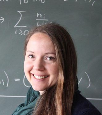 Das Foto zeigt das Institutsmitglied Roswitha Hofer. Sie hat brünette, lange Haare und tägt einen dunklebrünen Schal. Sie steht vor einer Tafel mit mathematischen Formeln.