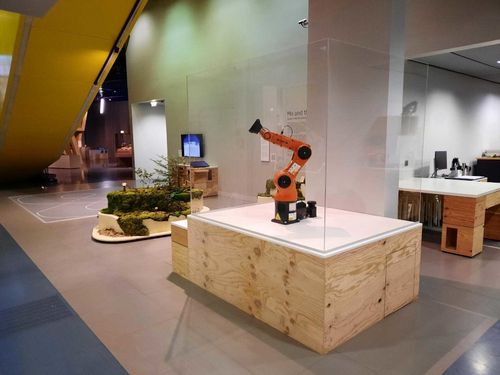 Blick in den Ausstellungsbereich: Im Vordergrund ist ein orangener Industrieroboterarm in einer Glasvitrine zu sehen, im Hintergrund eine kleine Insel mit Moos und Zweigen.