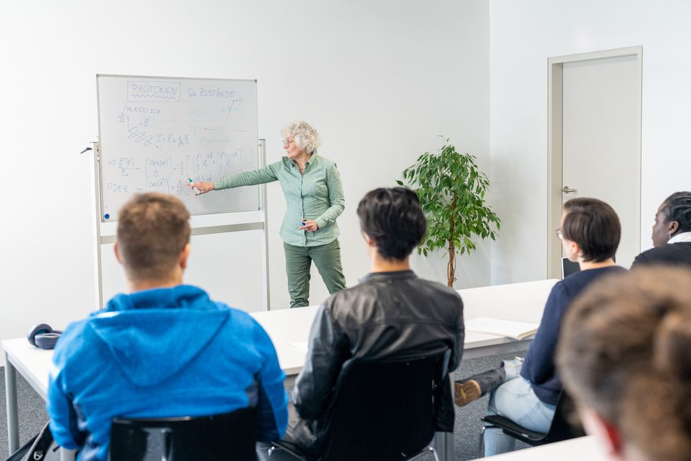 Ältere Lehrende mit weißen Haaren steht an der Tafel und unterrichtet eine Gruppe Student*innen