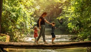 Bild einer Mutter, die, mit zwei Kindern an den Händen, auf einer schmalen Brücke einen Bach überquert.