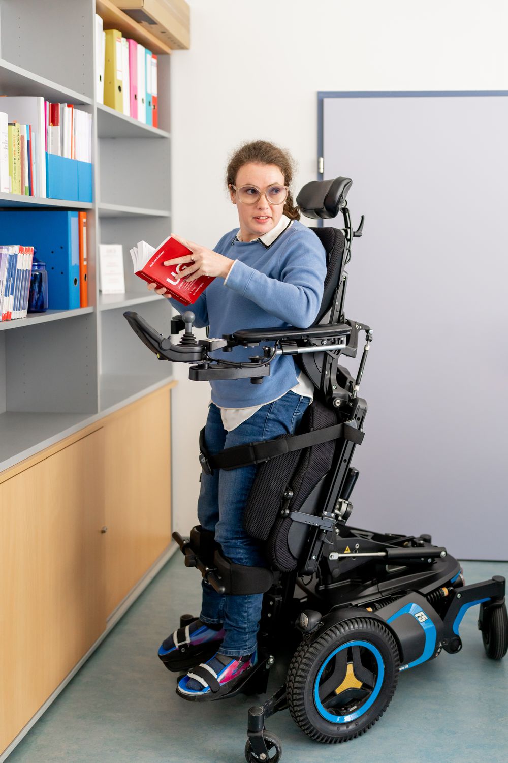 Mensch in modernem Rollstuhl mit Aufrichthilfe steht lesend vor Bücherwand