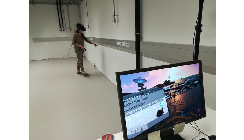 Schulterblick über Computerscreen auf dem eine VR-Spielwelt sichtbar ist, dahinter Person mit VR-Headset im Raum