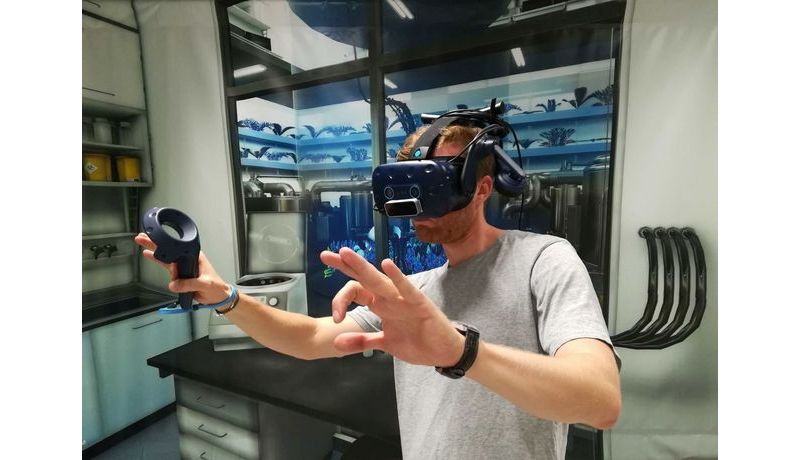 Mann mit VR-Brille und Controller in der Hand, im Hintergrund Plakatwand mit Laboransicht