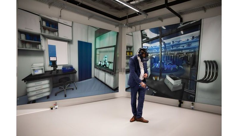 Mann mit VR-Brille und Controller in der Hand, im Hintergrund Plakate mit Laboransicht